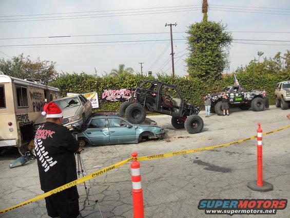 Several wrecked cars, a buggy, and a truck are in a lot with caution tape. A person in a Santa hat films