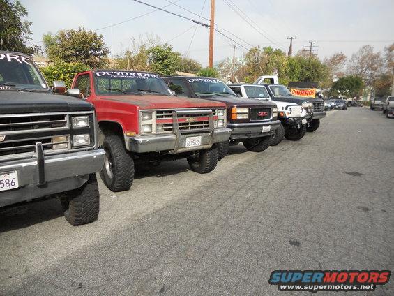 Several lifted pickup trucks parked in a row on a street. The red truck in front has a 'DEYMANS OFFROAD' banner on its windshield