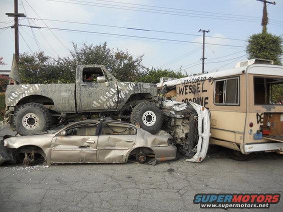 Mud-splattered pickup truck crushing a sedan, next to a bus with text