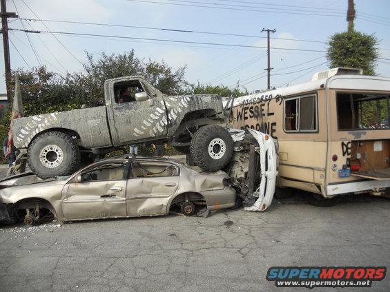 Wrecked pickup truck on top of a sedan, next to a bus. Tire tracks painted on truck