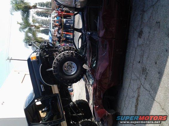 1986 Ford Bronco with large off-road tires and roll cage, next to a crushed car