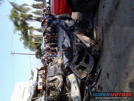 Heavily damaged car in a crowd, red truck visible