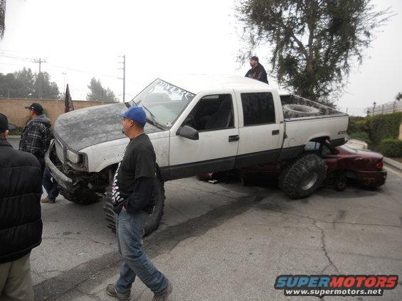Lifted white pickup truck with large tires is on top of a red sedan. People are standing around