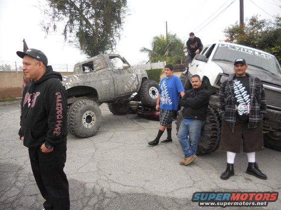 Four men stand around two heavily modified lifted trucks, one gray and one white