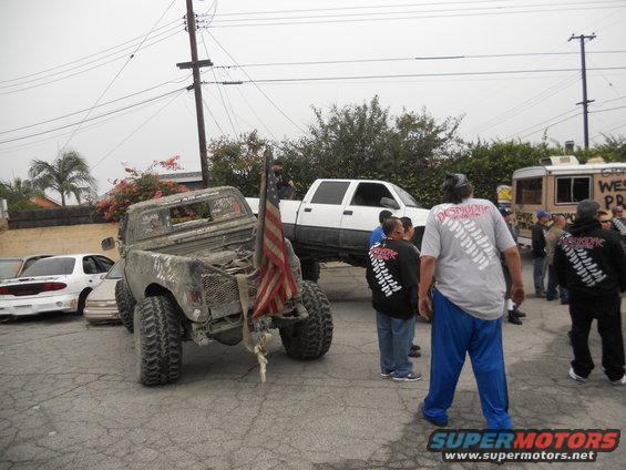 Mud-covered pickup truck with large tires and an American flag on the back, parked near a white crew cab truck and other people