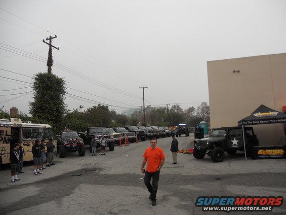 Several modified off-road vehicles and a van parked in a lot under an overcast sky