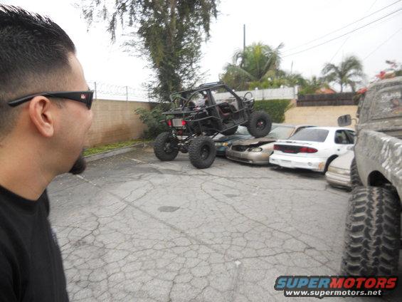 Off-road buggy perched on a pile of wrecked cars in a parking lot. A person looks on from the left