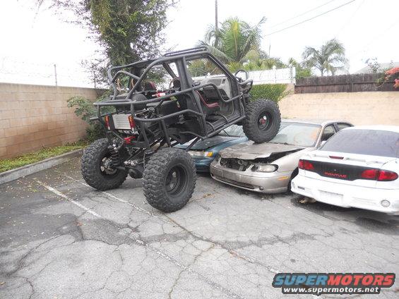 Custom off-road buggy with large tires is parked on top of three wrecked cars in a parking lot