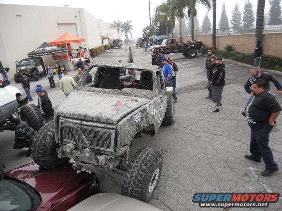 Mud-covered off-road truck with large tires, parked over a red car. Other vehicles and people are visible in the background