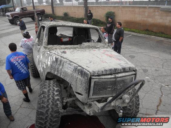 Mud-covered pickup truck with large tires, front end visible. Another brown pickup truck is parked in the background