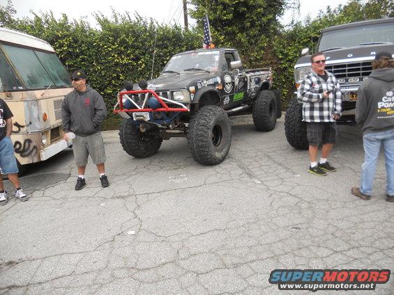 Black pickup truck with large tires and a red roll cage, parked next to a van and a Ford truck