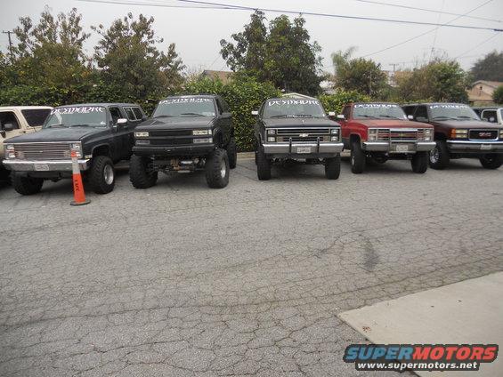 Several lifted trucks and SUVs parked in a lot. Some have banners on the windshields