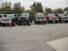 1986 Ford Bronco - A lineup of modified trucks and SUVs are parked on a cracked asphalt lot under an overcast sky.