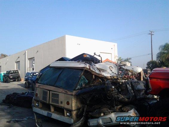 Front of a wrecked RV, heavily damaged, with other crushed vehicles in the background. A red truck is on the right