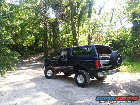 Dark blue Ford Bronco parked on a dirt road with trees in the background. Spare tire mounted on the back
