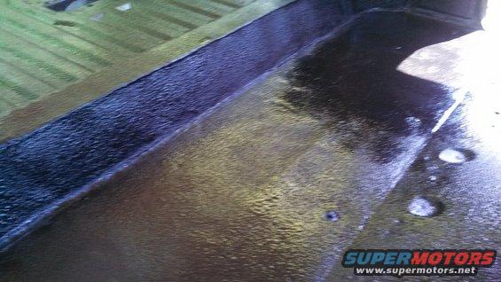 Close-up of a vehicle's interior floor with dark protective coating and some water droplets. Supermotors.net logo visible