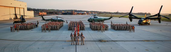 Military personnel in formation on an airfield with multiple helicopters and a large hangar