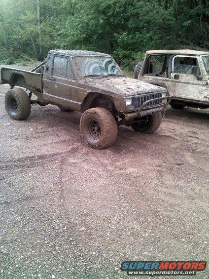 Mud-covered 1987 Jeep Comanche pickup truck with large tires, parked next to another muddy vehicle