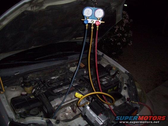 Car engine bay with AC manifold gauges connected to the system. A vacuum pump is also attached