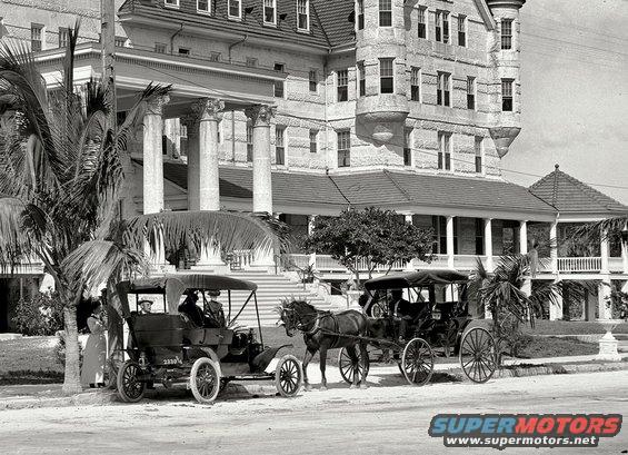 Black 1909 Ford Model T with a black top, parked on a street next to a horse-drawn carriage
