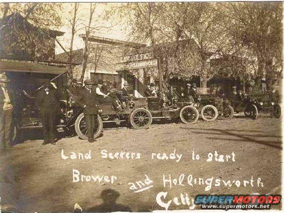 Several early automobiles parked on a dirt road in front of buildings, with people standing around them. Text reads 'Land Seekers ready to start Brower and Hollingsworth Celly'