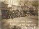 1909 Ford Model T - Multiple early automobiles are lined up on a dirt road, with people gathered around them. Text indicates a 'Land Seekers' event.