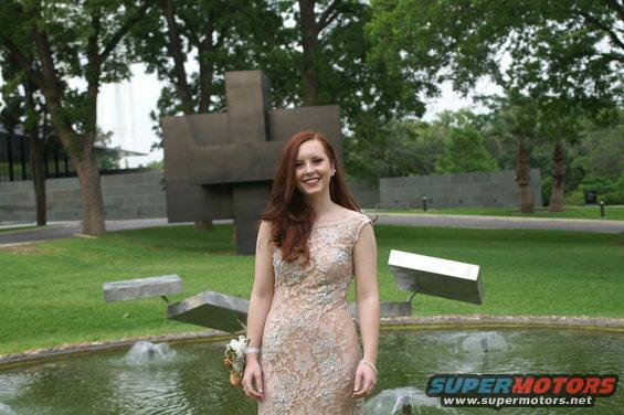 Woman in a light-colored, beaded formal dress stands in front of a fountain and modern sculpture