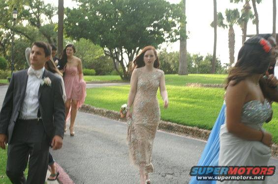 People in formal attire walking on a paved path outdoors with trees and grass in the background