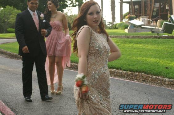 Three young people in formal attire on a paved path with grass and buildings in the background