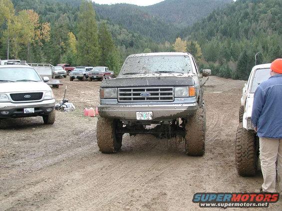 Muddy 1988 Ford Bronco with lifted suspension and large tires, parked on a dirt track. Another SUV is to the left, and a person in an orange hat is to the right