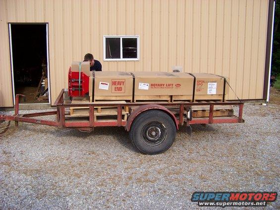 Red trailer loaded with cardboard boxes and a red piece of equipment, parked on gravel in front of a tan building