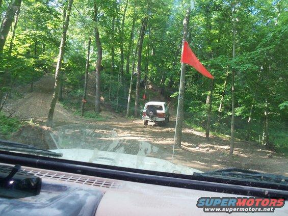 White 1989 Ford Bronco seen from behind on a dirt trail in a forest, with a red flag on a pole