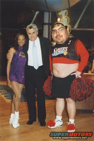 Jay Leno poses with a woman in a purple Lakers outfit and a person in a Philadelphia 76ers jersey and skirt holding pom-poms