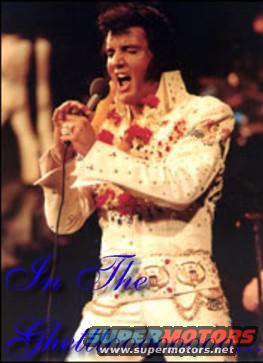 Elvis Presley in a white jumpsuit with floral lei, singing into a microphone on stage