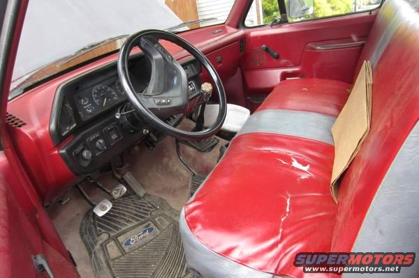 Interior view of a red and gray bench seat, dashboard, and steering wheel in a pickup truck. Ford floor mat visible