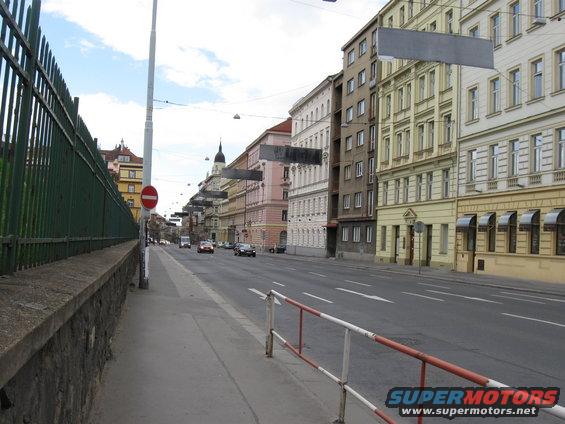 Street view with buildings, cars, and a green fence