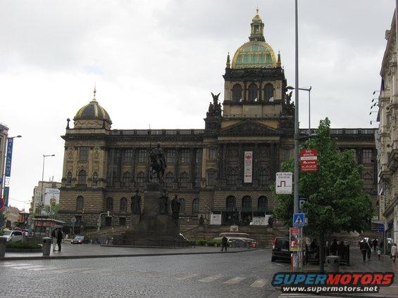 Historic building with a dome and a statue in a city square, with a few cars and pedestrians visible