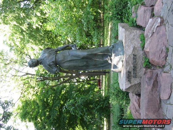 Statue of a woman in a long dress standing next to a large rock with text on it, in a park setting