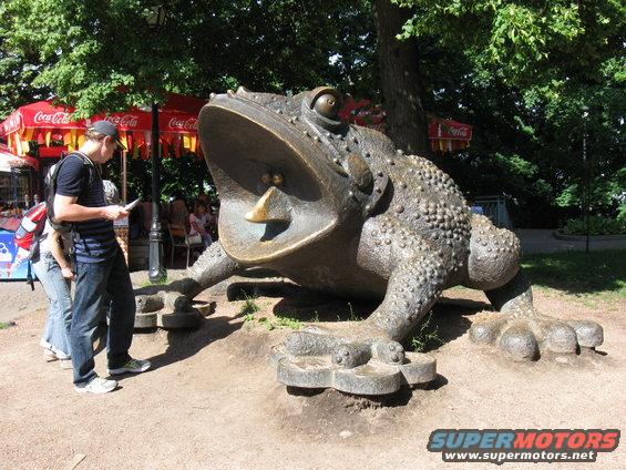 Bronze toad sculpture with open mouth, people nearby, Coca-Cola umbrellas in background