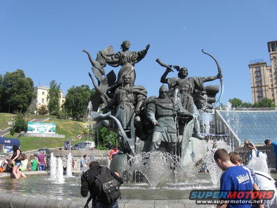 Monumental fountain with bronze sculptures of warriors and figures, with water spraying. People are visible in the foreground and background