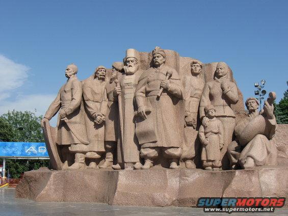 Stone monument depicting a group of people in historical attire, with a blue sign in the background