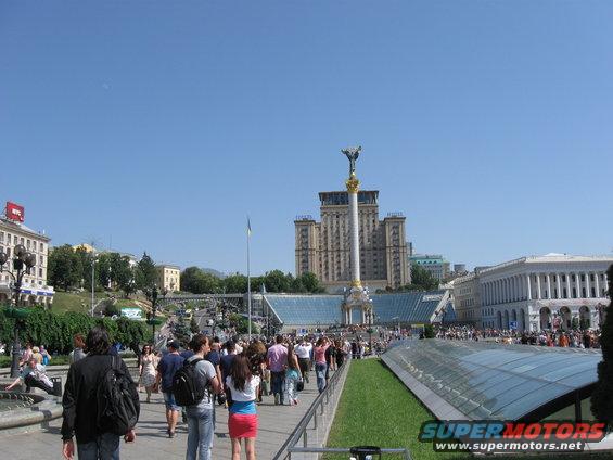 City square with a tall monument topped by a statue, surrounded by buildings and a crowd of people walking