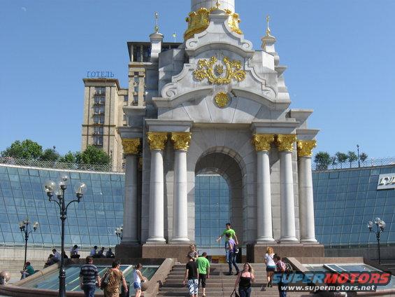 Monumental archway with golden accents and columns, people walking on stairs below. Hotel building in background