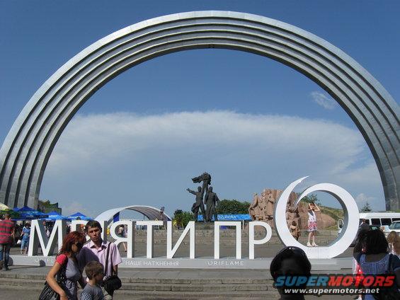 Giant metal arch over sculptures and large white letters spelling 'МРІЯТИ ПР'. People are standing around