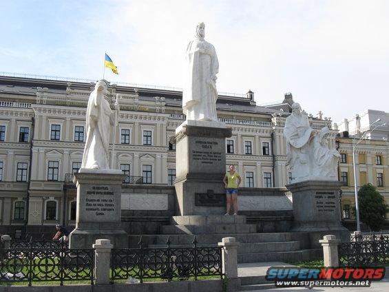 Monument with white statues and a Ukrainian flag, in front of a light-colored building. A person stands on the monument's steps