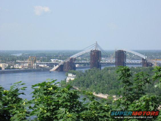 Bridge under construction over a wide river, with trees in the foreground