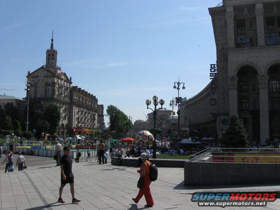 City square with people walking, large buildings, and a giant soccer ball decoration