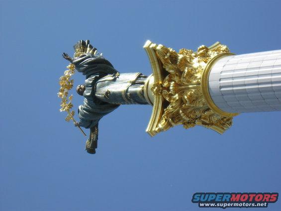 Statue holding a golden wreath atop a decorative column against a blue sky. Supermotors.net logo in lower right