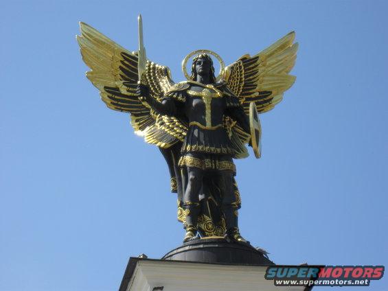 Statue of an angel holding a sword and shield against a blue sky. Gold wings and trim