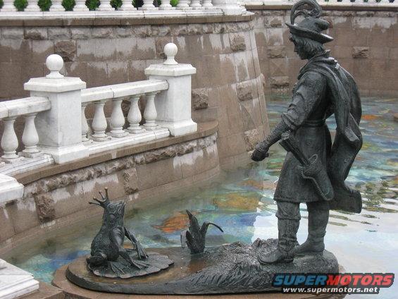 Bronze statue of a man with a bow and quiver facing a frog wearing a crown, in a fountain